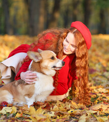 A girl with long red hair, in a red coat and beret, plays with her corgi dog, in the park, in the fall. Golden autumn, walk with the dog, friendship. Beautiful autumn image