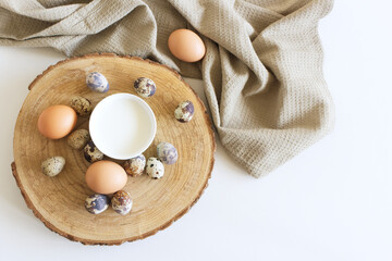 Chicken eggs brown and Quail eggs, on a cut wooden tree trunk on a kitchen towel on a white table and a cup of milk. Concept farm products and natural nutrition.