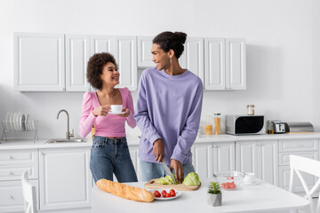 Happy african american woman with cup looking at boyfriend cooking salad in kitchen.