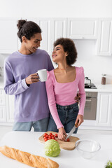 Smiling man holding cup and hugging african amerian girlfriend cutting cherry tomato at home.