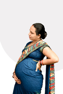 An Indian pregnant lady holding her left back and lower belly with one hand eyes closed feeling, side view photo having light grey and white background selectively focused