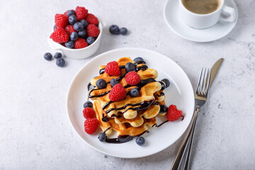 Viennese or Belgian waffles with fresh berries (raspberries and blueberries) and chocolate sauce on a white plate and a cup of coffee. Traditional dessert. Close-up, white background.