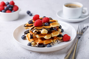 Viennese or Belgian waffles with fresh berries (raspberries and blueberries) and chocolate sauce on a white plate and a cup of coffee. Traditional dessert. Close-up, white background, side view.