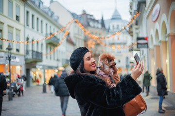 Happy emotional young dark-haired woman in a faux fur coat and beret makes a selfie with her dog apricot little poodle on the street of a European city. Winter holidays.