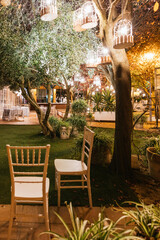 Illuminated night terrace with two chairs in the center, a tree in the central lawn and chandeliers hanging from the branches