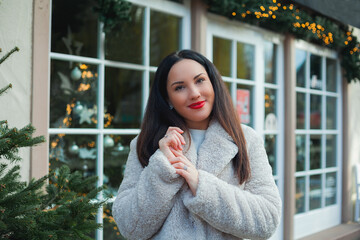 Christmas concept. Beautiful young dark-haired woman in a beige eco-coat on the street of the Christmas city. Stylish model is shopping in a European city during the winter break. New Year.