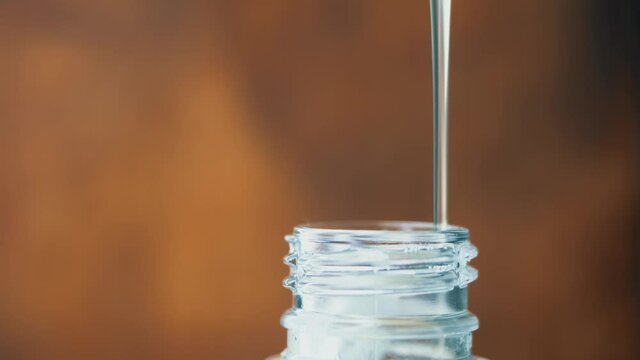 Thick transparent liquid slowly pours in thin stream. Plastic bottle neck. Syrup spreads along thread. Liquid soap or gel dish drips into container. Close-up side. Unknown chemical substance is poured
