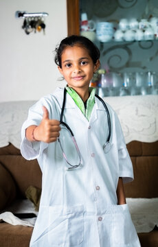 Portrait shot of Happy smiling girl with Doctor dress and stethoscope showing thumbs up at home - concept of childhod dreams, future planning and career imagination