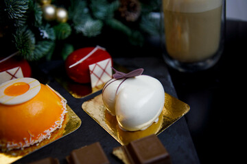 a table with sweets (mousse cakes of various shapes and colors) and pieces of chocolate. Spruce branches decorated for Christmas are visible in the background