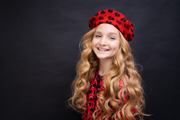 Following her personal style. Little girl in a french style hat. Happy girl with long curly hair in a red beret and dress. Parisian child on a black background. Summer fashion and beauty.