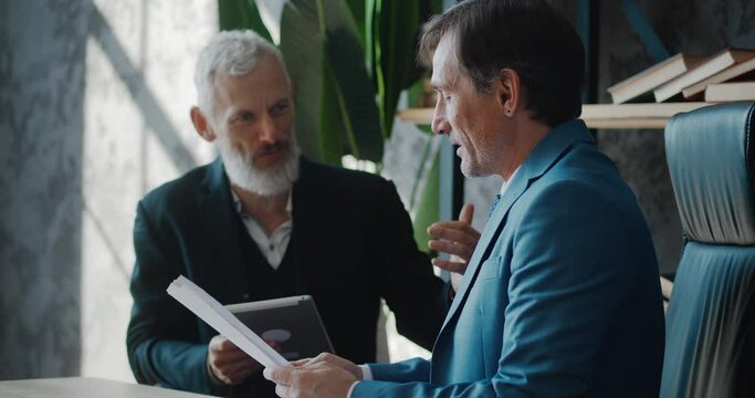 Two mature businessmen discussing business growth in modern office while sitting at desk. Man ceo talking with colleague about deal while meeting indoors