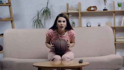 astonished sport fan woman sitting on couch with open mouth while watching basketball match.