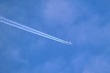Distant passenger jet plane flying on high altitude through white clouds on blue sky leaving white smoke trace of contrail behind. Air transportation concept