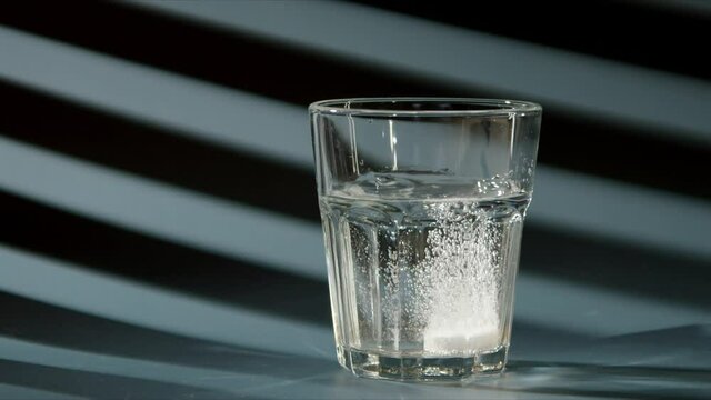 Effervescent tablet falls into a glass with clear clear water. Soluble medicine in water. blue background, sunlight shines through blinds. Slow motion video of medical drink.