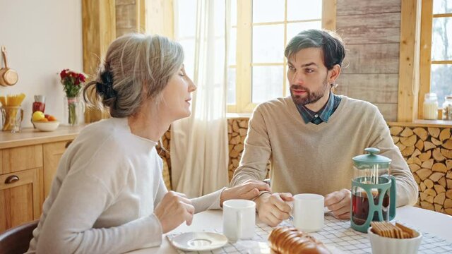 Young upset man telling his problems to mom, caring grey haired woman calming him, talking together at home