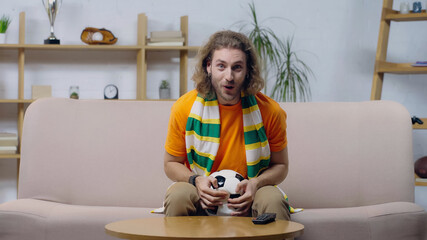 amazed sport fan in striped scarf sitting on couch with ball and watching football championship.