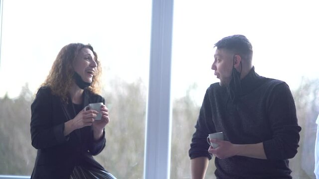 Colleagues talk during break in office kitchen. Curly woman communicates with workmate hold coffee cups enjoy pleasant conversation, discuss project. Good relations at work concept