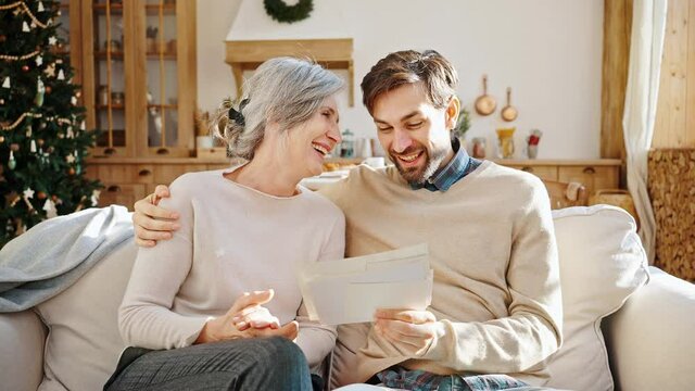 Young positive man showing his elderly mother photos of grandchildren, sitting together at home on Christmas holidays