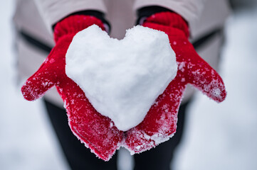 a heart made of snow in the hands of a girl in red mittens on a background of snow in winter. High quality photo