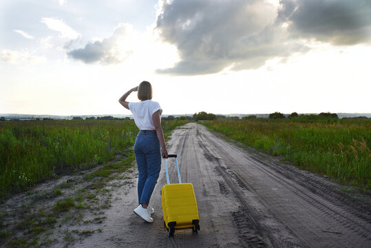 a girl with a small yellow suitcase walks along an empty country road on a summer day