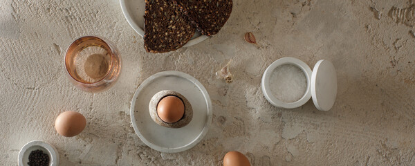 Obraz na płótnie Canvas Breakfast setting with boiled egg in stone egg cup, whole grain rye bread with seeds, glass of water, salt flakes and pepper in concrete bowls on textured clay background. Healthy breakfast concept.