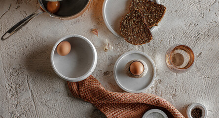 Breakfast setting with boiled egg in stone egg cup, whole grain rye bread with seeds, glass of water, salt flakes and pepper in concrete bowls on textured clay background. Healthy breakfast concept.