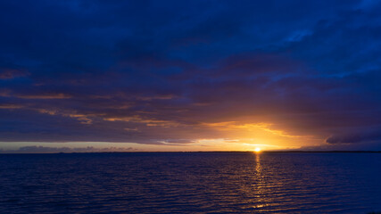 The sun sets as a golden circle touching the horizon between low hanging clouds and the sea on the Rømø causeway in Denmark