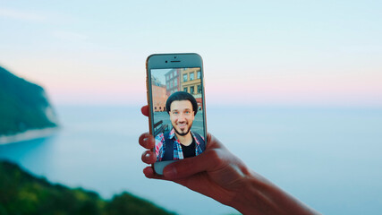 Close-up of hand holding smartphone during video call with man in front of the sea. Beautiful landscape in the background.