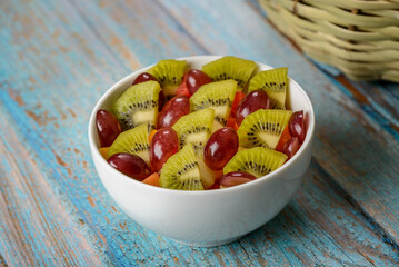 Fruit salad with grape, kiwi and mango in white bowl on blue wooden table.
