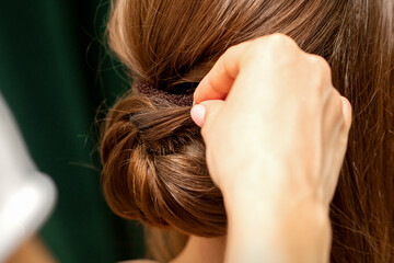 Hands of hairdresser making french twist hairstyle of an unrecognizable young brunette woman in a beauty salon, back view, close up