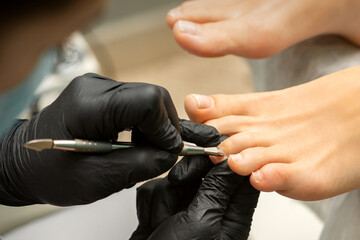 Cuticle Removal on Toes. Hands in black gloves of pedicure master remove cuticle on female toes by pusher - obrazy, fototapety, plakaty
