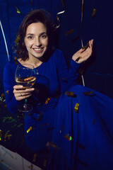 cheerful Caucasian brunette girl in a blue dress with a glass of champagne is sitting on sofa in a restaurant with confetti on a holiday
