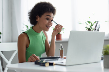 Smiling african american woman applying eye shadow near cosmetics and blurred laptop at home.