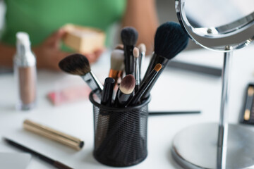 Cropped view of cosmetic brushes near mirror and blurred african american woman.