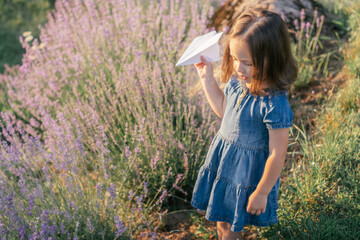 Little girl 3-4 with dark hair in denim dress in sun launches paper plane among large bushes of lilac lavender