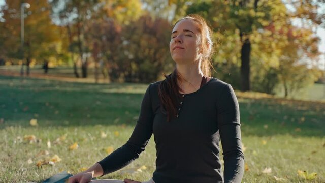 Young woman having a meditation sitting on mat in lotus position in city park. Sunny autumn day in a city park. Sporty girl practicing meditation outdoors. High quality 4k footage