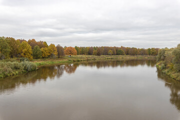 Autumn landscape with a river. Cloudy autumn day by the river. View of the river with trees and bushes on the bank. Bright colors of autumn on a cloudy day.