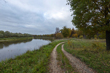 Fototapeta na wymiar Dirt road along the river bank. Cloudy autumn weather. Rural landscape with road, river and autumn forest.