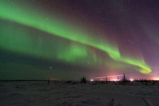 The northern lights and aurora borealis fill the sky above distant city lights near Churchill, Manitoba, Canada