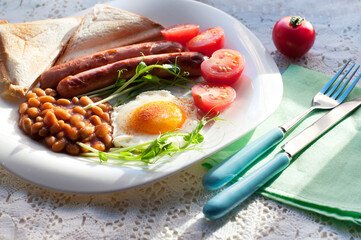 English breakfast with scrambled egg, sausages, toast, tomatoes, pea sprouts and beans on white plate. Traditional English breakfast with fork and knife on white lace tablecloth