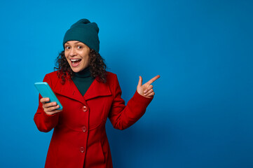 Pensive awesome happy cheerful woman wearing bright red coat and warm green woolen hat holding smartphone and rejoices pointing with her finger on a copy space for ads, isolated on blue background