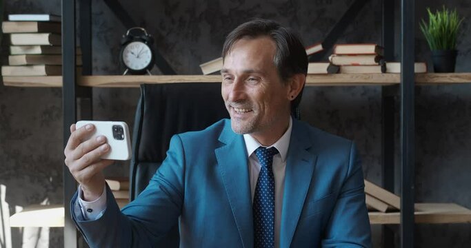 Happy mature businessman making facetime video calling with smartphone at modern office, greeting people on phone screen. Male boss in suit making selfie video chat online on mobile phone at workplace