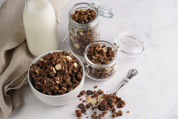 Breakfast muesli cereal with chocolate chips and nuts in several jars and bowl, bottle of milk, white surface