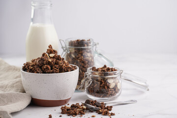 Breakfast muesli cereal with chocolate chips and nuts in several jars and bowl, bottle of milk, white surface