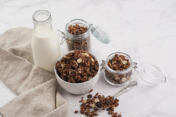 Breakfast muesli cereal with chocolate chips and nuts in several jars and bowl, bottle of milk, white surface
