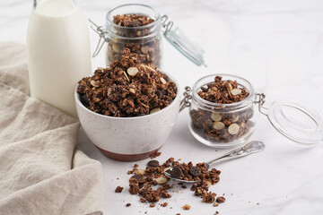 Breakfast muesli cereal with chocolate chips and nuts in several jars and bowl, bottle of milk, white surface