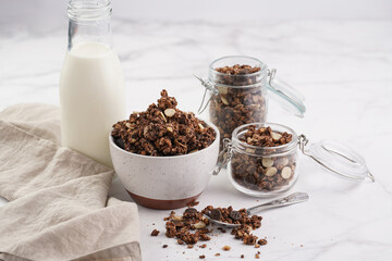 Breakfast muesli cereal with chocolate chips and nuts in several jars and bowl, bottle of milk, white surface