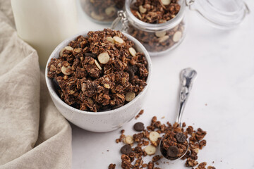 Breakfast muesli cereal with chocolate chips and nuts in several jars and bowl, bottle of milk, white surface