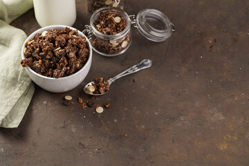 Breakfast muesli cereal with chocolate chips and nuts in several jars and bowl, bottle of milk, brown wooden surface