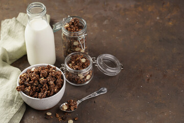 Breakfast muesli cereal with chocolate chips and nuts in several jars and bowl, bottle of milk, brown wooden surface